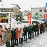 大雪の中で衆議院議員総選挙の期日前投票所に並ぶ有権者の長い行列。自民党の高市早苗氏の選挙ポスターや幟(のぼり)が並び、背景の電光掲示板には「投票率56.26%」「高市旋風」の文字が表示されている冬の選挙風景。