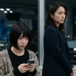 夜のオフィスで深刻な表情を浮かべスマートフォンで情報を確認する女性と、背後で見守るスーツ姿の女性