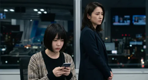 夜のオフィスで深刻な表情を浮かべスマートフォンで情報を確認する女性と、背後で見守るスーツ姿の女性